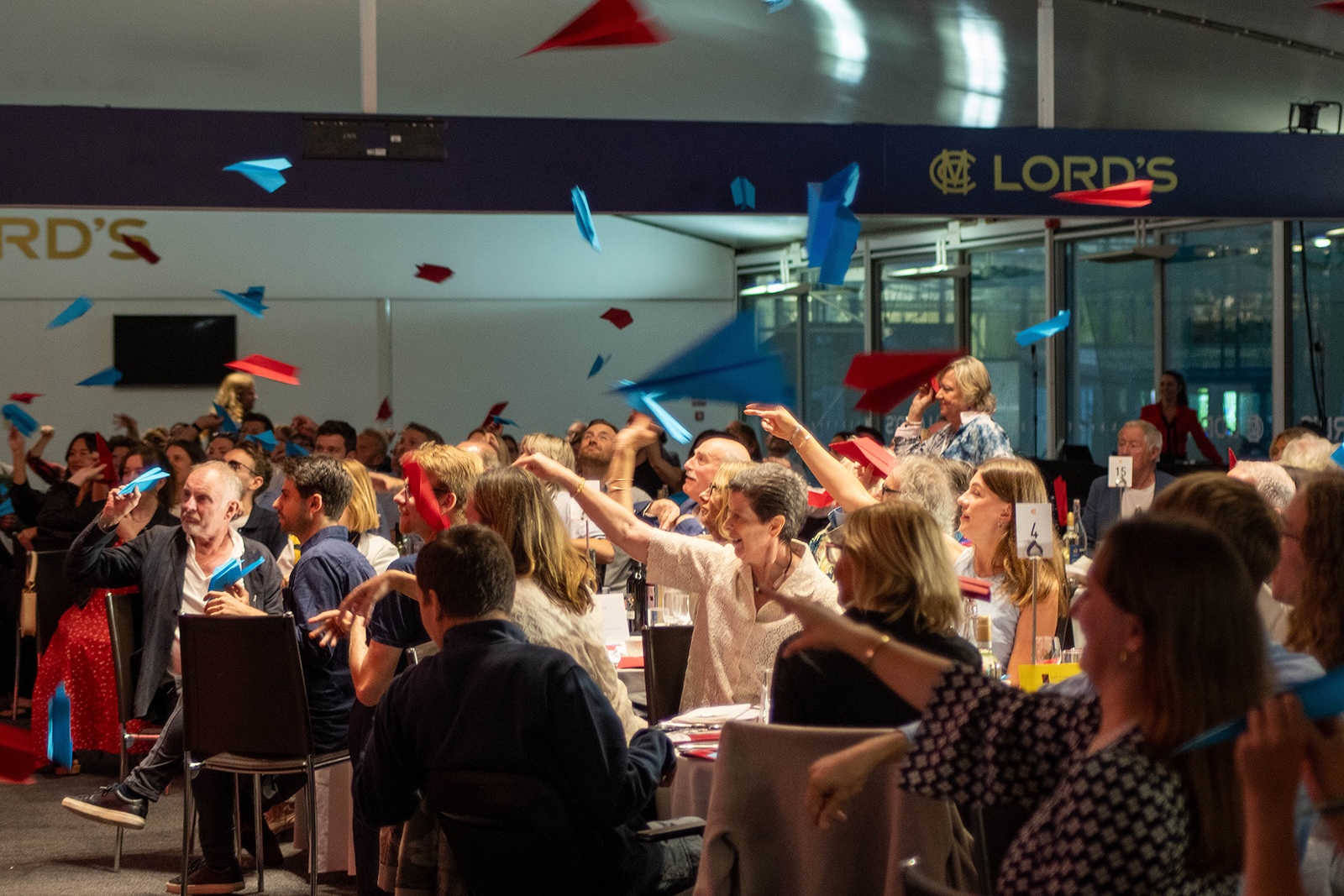 Crowd of people in the Nursery Pavilion at Lord's throwing red and blue paper aeroplanes towards the stage on the left hand side.jpg