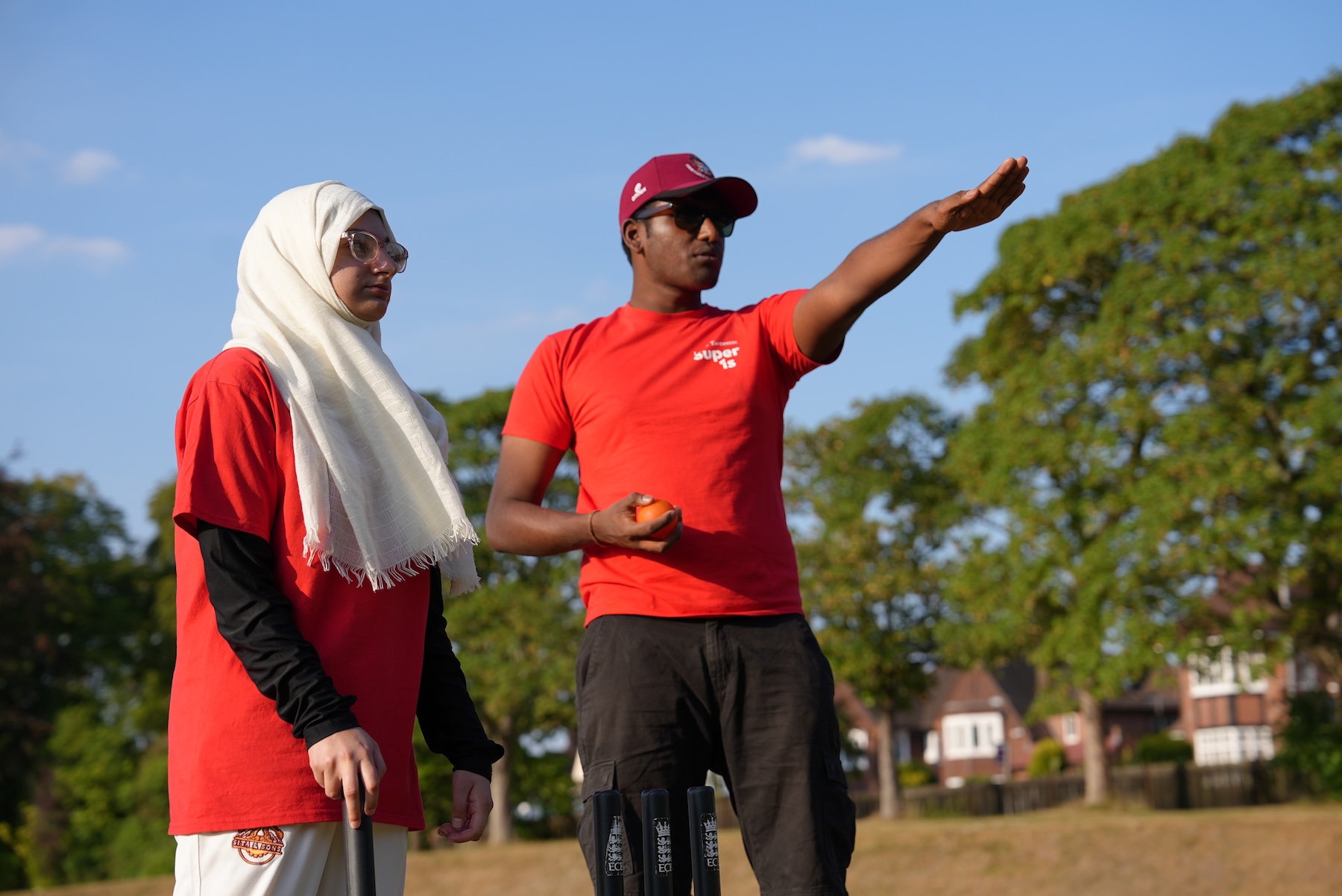 Prajith in a red Super 1s t-shirt with his forearm outstretched ready to bowl with a girl standing next to him with a bat in her hand