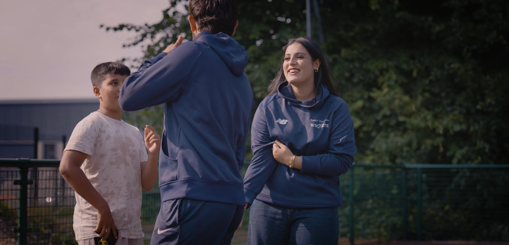 Maha smiling with another coach who has their back turned to the camera, with a young person stood in the background on the left.png