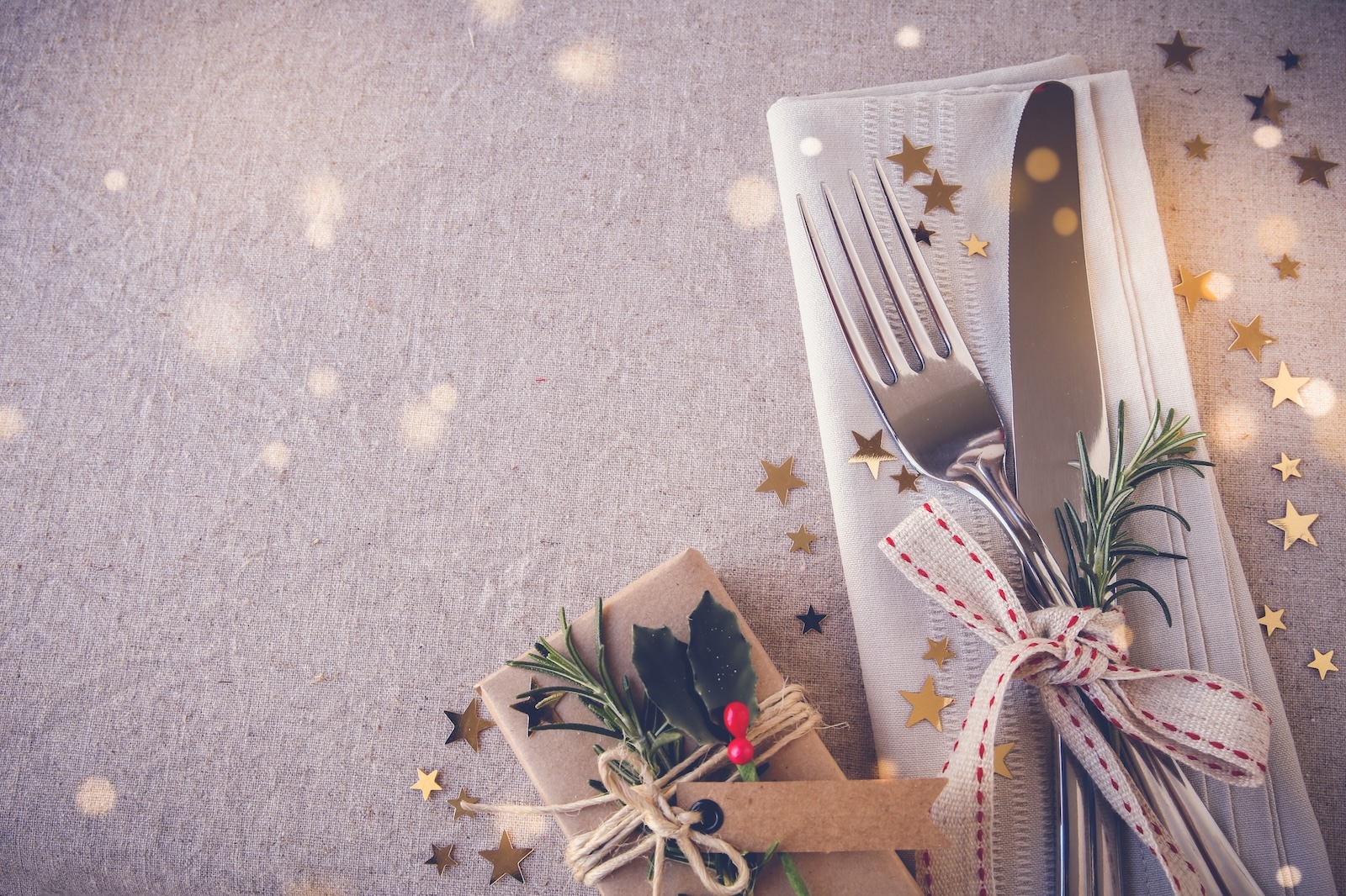 Fork and knife on napkin tied together with ribbon nect to a brown parcel tied with string and a spring of holly. Gold stars sprinkled around table next to the items.jpeg